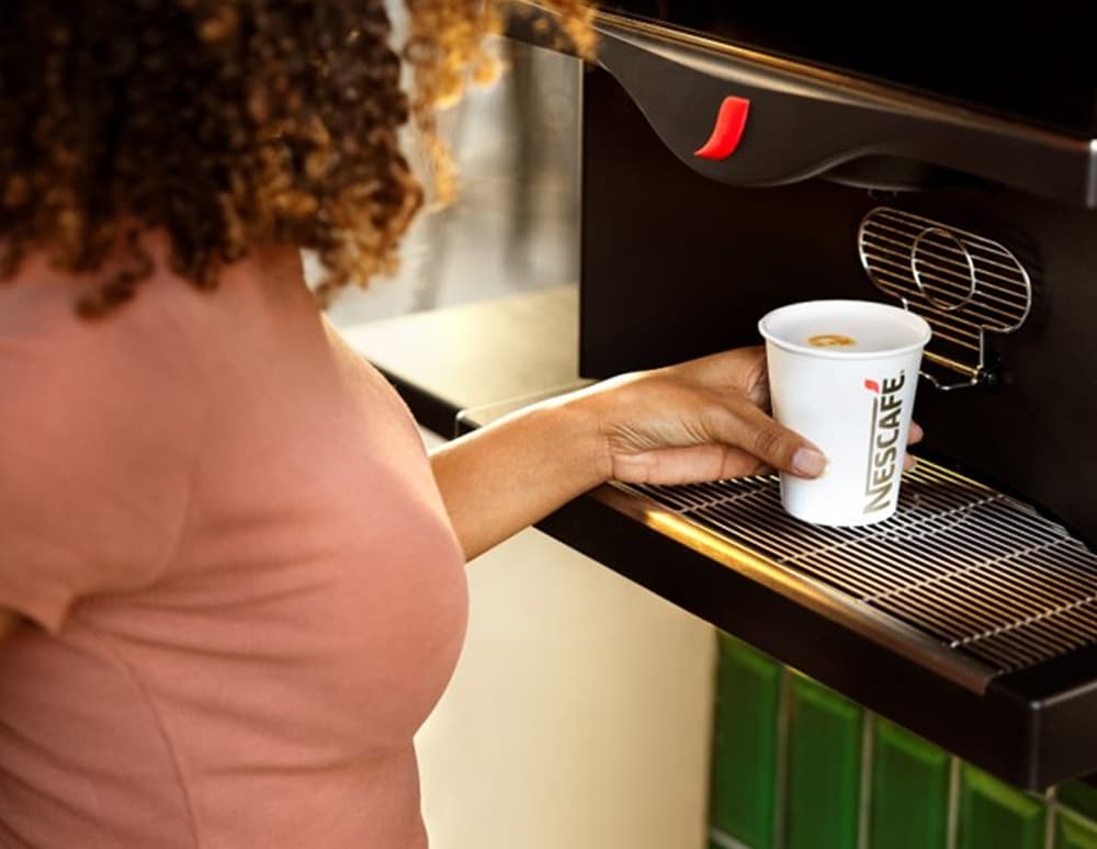 Une femme aux cheveux bouclés tient une tasse Nescafé et se sert un café dans une machine à café élégante.