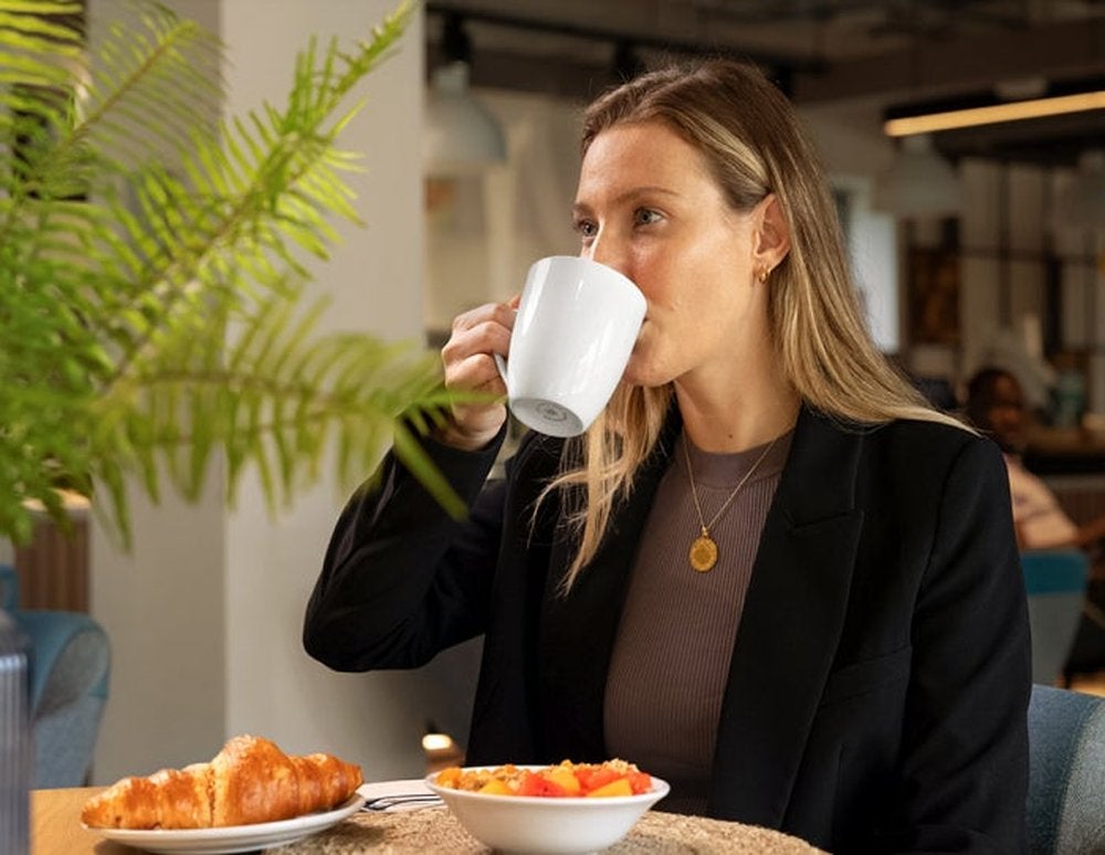 A woman in a black blazer sips from a white mug, with a plate of croissants and fruit salad on the table