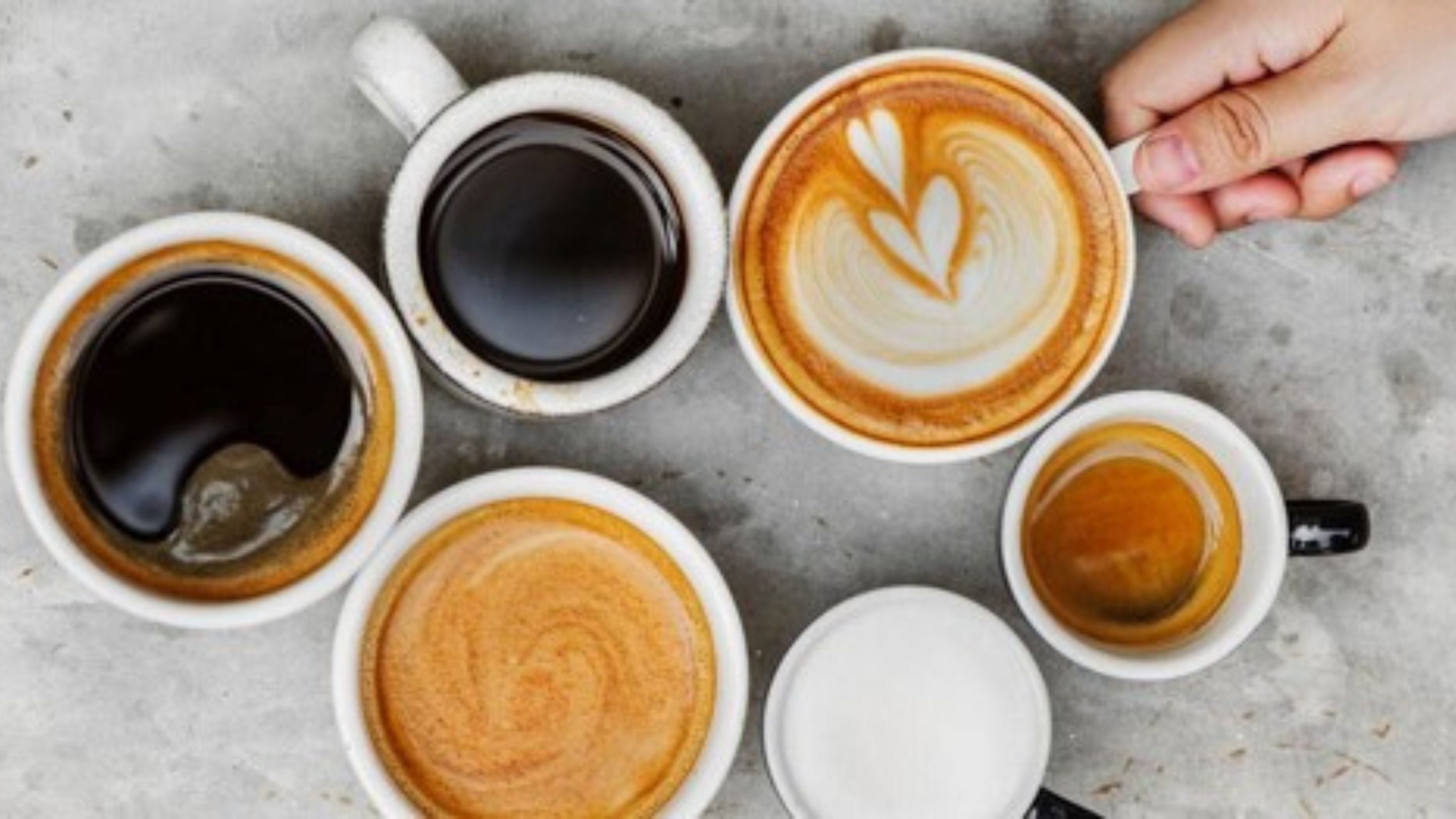 An assortment of coffee cups, including espresso, black coffee, and latte art, with a hand holding one cup