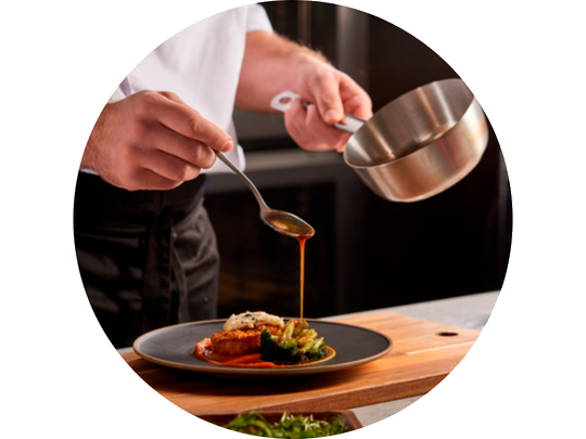 A chef drizzles sauce over a plated dish of meat and vegetables on a wooden cutting board in a kitchen setting.