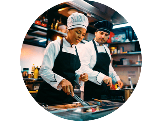 Two chefs in professional attire prepare food on a grill in a well-stocked kitchen, focused on their culinary tasks.