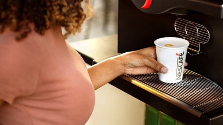 Une femme aux cheveux bouclés tient une tasse Nescafé et se sert un café dans une machine à café élégante.