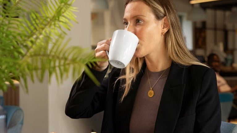A woman in a black blazer sips from a white mug, with a plate of croissants and fruit salad on the table