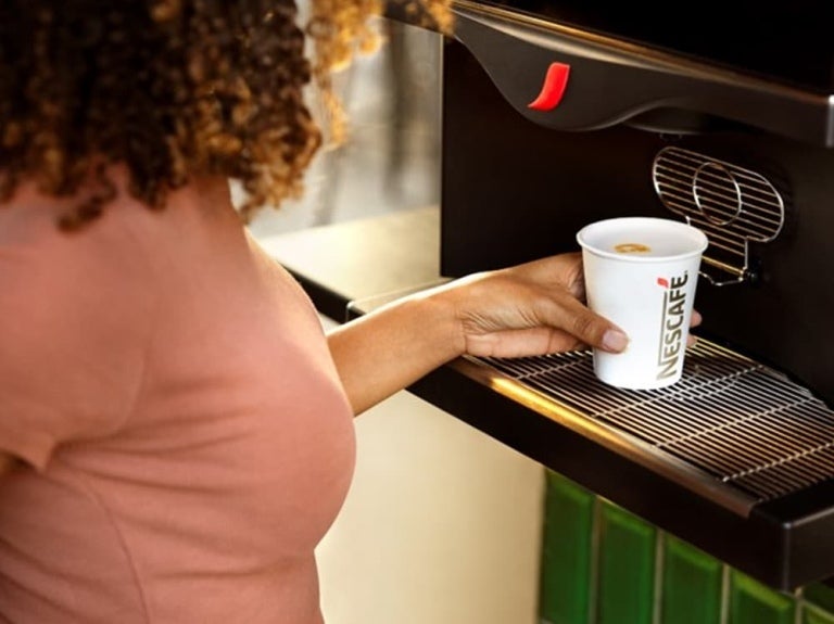 Une femme aux cheveux bouclés tient une tasse Nescafé et se sert un café dans une machine à café élégante.
