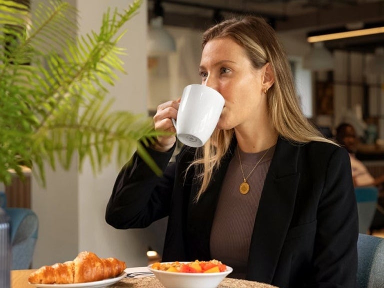 A woman in a black blazer sips from a white mug, with a plate of croissants and fruit salad on the table