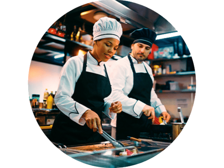 Two chefs in professional attire prepare food on a grill in a well-stocked kitchen, focused on their culinary tasks.