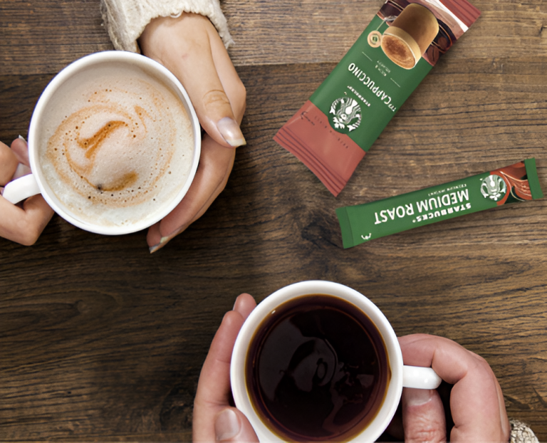 Two people holding mugs of coffee on a wooden table with Starbucks Cappuccino and Medium Roast packets nearby.